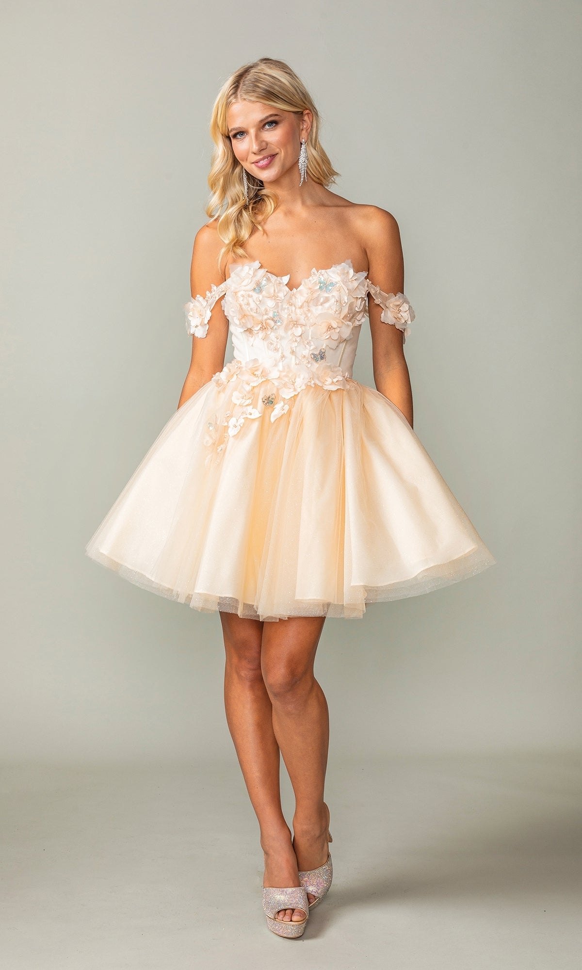 A woman stands smiling in the Off-Shoulder Short Babydoll Homecoming Dress 3388 with floral appliqués and high heels, set against a plain light background.